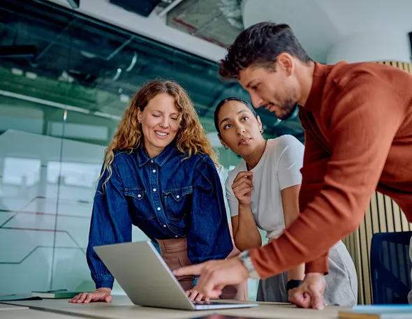 Three colleagues standing around a laptop in a glass-walled office, one pointing at the screen