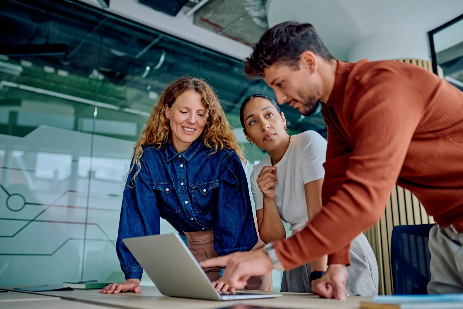 Three colleagues standing around a laptop in a glass-walled office, one pointing at the screen