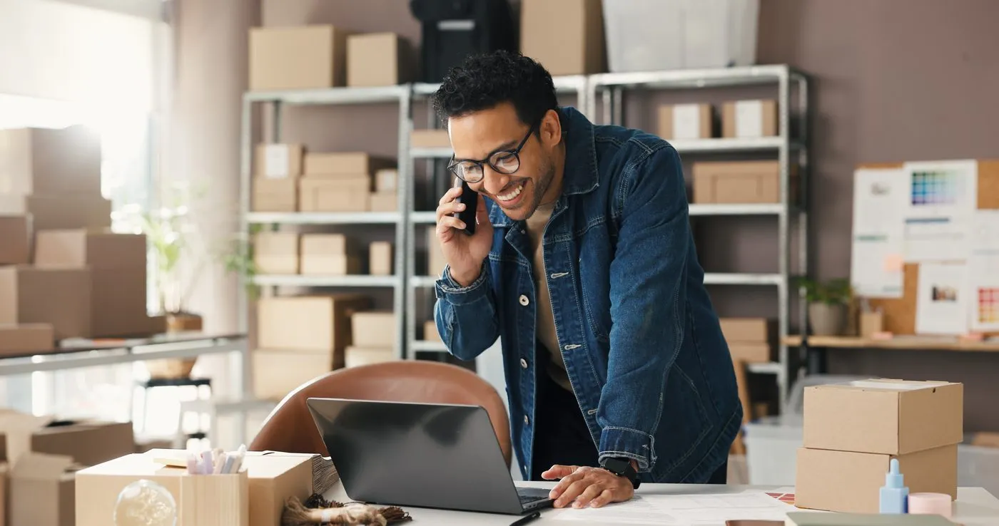 A business owner on the phone at a laptop, surrounded by stacked parcels
