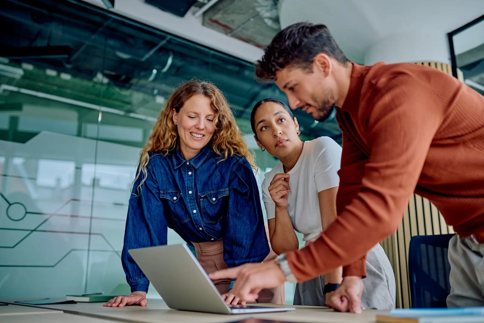 Three colleagues gathered around a laptop in a glass-walled office