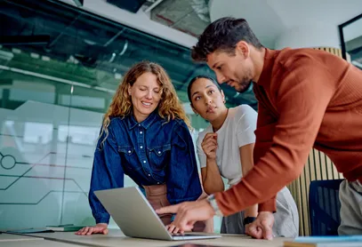 Three colleagues gathered around a laptop in a glass-walled office