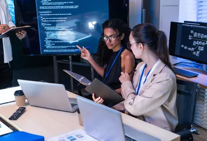 Two colleagues at a bank of monitors, one pointing at code on a large screen